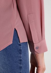 Blouse rose clair texturée à manches longues, avec poignets boutonnés et fentes latérales, associée à un jean bleu.