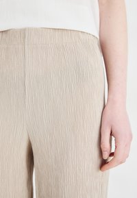 Lys beige, teksturerte bukser med et krøllete design. Elastisk midjebånd for komfort. Nærbilde av en hånd som hviler på stoffet.