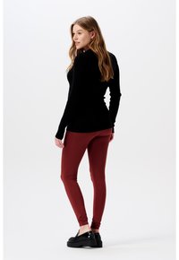 Zwarte geribbelde top met lange mouwen in combinatie met bordeauxrode leggings. Het model draagt zwarte platform schoenen, haar in losse golven, tegen een effen achtergrond.