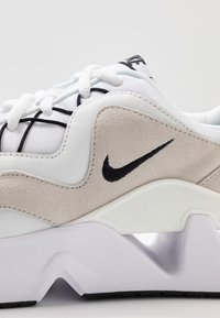 Valkoinen lenkkari, jossa on teksturoitu harmaa mokka-akseli, mustat nauhat ja musta Nike-logomerkki. Varusteena on paksu valkoinen pohja, jossa on aaltoilukuviota.