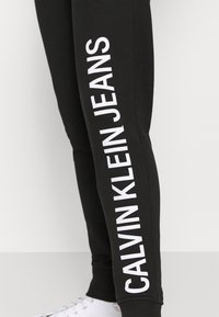 Černé jogger kalhoty s bílým textem "CALVIN KLEIN JEANS" po straně, vyrobené z měkké tkaniny s přiléhavým střihem a elastickým pasem.