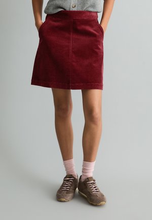 Person trägt einen roten Cordrock mit den Händen in den Taschen, pinke Socken, braune Sneakers und ein graues Oberteil vor neutralem Hintergrund.