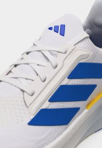 Λευκό αθλητικό παπούτσι με μπλε ρίγες και λογότυπο, λευκά κορδόνια και κίτρινη λεπτομέρεια στη σόλα, εμφανιζόμενο σε κοντινό πλάνο.