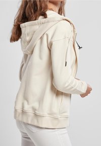 Krémová zipová mikina z měkké tkaniny, s kapucí, žebrovanými manžetami a lemem, a bočním zipovým kapsou. Hladký, uvolněný střih.