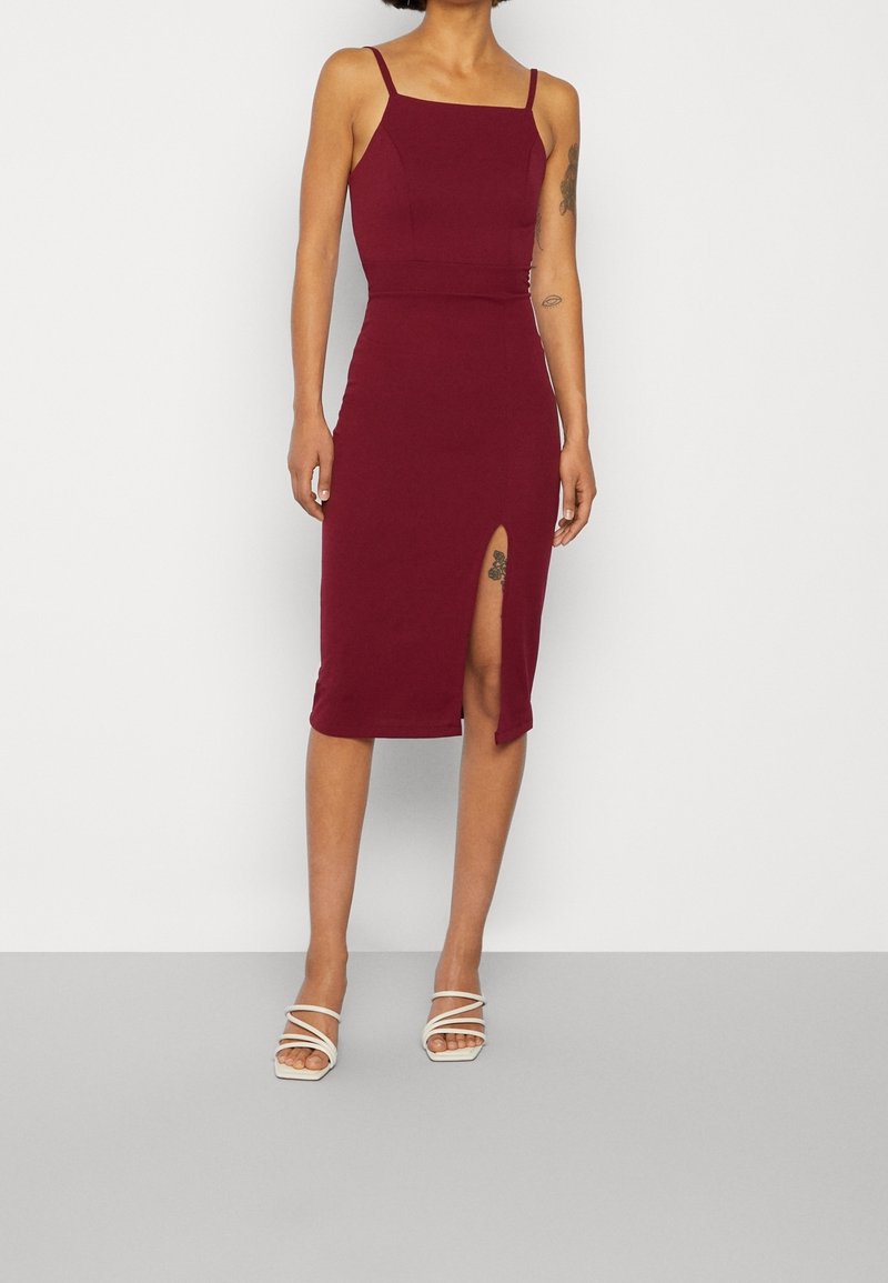 Femme portant une robe midi bordeaux ajustée sans manches avec une fente à la cuisse et des talons blancs à brides, debout devant un fond uni.