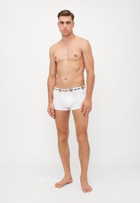 Weiße Baumwoll-Boxer-Shorts mit einem schwarzen Moschino-Logo-Bund, auf einem Kleidungsständer vor einem neutralen Hintergrund präsentiert.