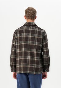 Man met kort, krullend haar, gekleed in een bruine en grijze geruite jas en een blauwe broek, staand met zijn gezicht naar een witte achtergrond.