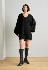 Vestido negro con una apariencia texturizada y esponjosa, mangas anchas y escote en V. Combinado con botas de cuero negro hasta la rodilla con tacones de bloque.
