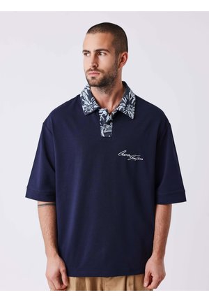 Homme aux cheveux courts et à la barbe portant une chemise bleu marine à manches courtes avec un col à motifs et un pantalon beige, regardant légèrement sur le côté.