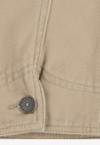 Beige Canvasstoff mit einer genähten Naht und einem Metallknopf mit eingraviertem Markennamen, der eine strapazierfähige und strukturierte Oberfläche zeigt.