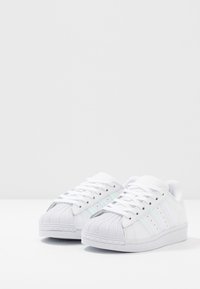 Vita sneakers med en texturerad gummitå, tre iriserande ränder på sidorna och en slät ovandel. Snörningsdesign med en platt sula.