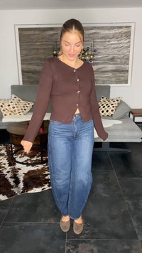 Cardigã botão castanho de manga longa com uma cintura ajustada e decote em V, combinado com jeans azul de perna larga e sapatilhas cinzas.