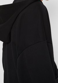 Sudadera negra con capucha que presenta una textura suave, un corte relajado y hombros caídos. Diseño minimalista sin logotipos ni patrones visibles.