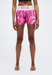 Γυναίκα που φοράει λαμπερά ροζ αθλητικά σορτς με τη μάρκα Venum και μαύρο αθλητικό σουτιέν, όρθια, ξυπόλητη, μπροστά σε λευκό φόντο.