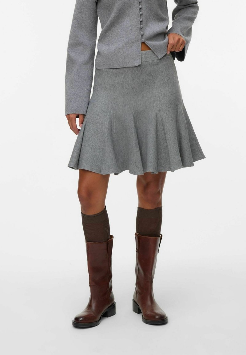 Femme portant une jupe plissée grise, une veste grise boutonnée assortie, des chaussettes hautes foncées et des bottes en cuir marron, debout sur un fond blanc.