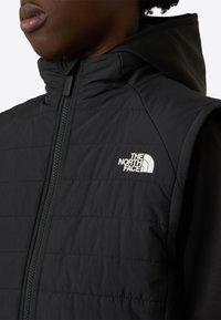 Čierna zateplená vesta s prešívaným dizajnom a vysokým golierom, s predným zipsom a logom The North Face v bielej farbe na hrudi.