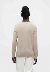 Bež dolgo rokav pulover iz mehkega materiala, z enostavnim dizajnom in rebrastimi manšetami, prikazan od zadaj na svetli podlagi.