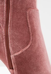 Gros plan sur une botte en velours rose poussiéreux avec une fermeture éclair latérale visible et des détails de panneaux cousus.