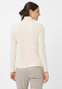 Frau mit dunklem Haar im Dutt, die ein cremefarbenes Langarmoberteil und gemusterte beige, schwarze und weiße Karohosen trägt, leicht zur Seite gedreht.