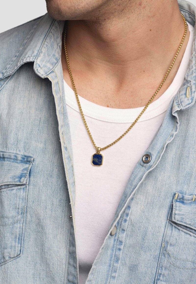 Guldkæde med en blå pendants i hexagonal form, båret over en hvid skjorte under en lyseblå denimjakke.