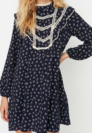 Frau trägt ein marineblaues langärmeliges Kleid mit weißen Blumenmustern und beigen Spitzenakzenten im Brust- und Schulterbereich.