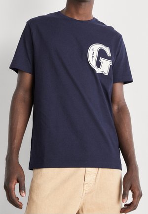 T-shirt en coton bleu marine à manches courtes avec un grand logo "GANT" blanc sur le côté gauche de la poitrine. Tissu léger et respirant.