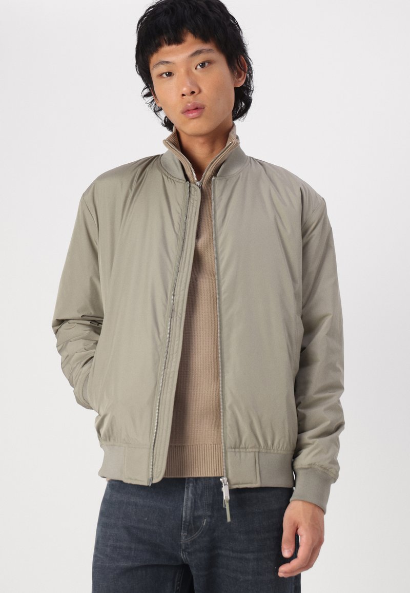 Lichtbeige bomberjack met een opstaande kraag, ritsluiting en elastische manchetten. Draagt over een beige gebreide trui en donkere jeans.