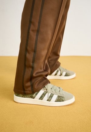 Baskets camouflées vertes avec des rayures blanches, en tissu texturé et semelle en caoutchouc, associées à un pantalon de jogging marron avec des rayures latérales foncées.