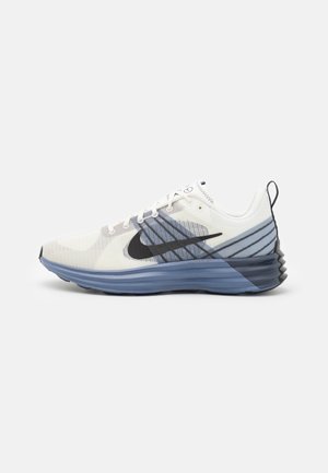 Biało-niebieski but sportowy z czarnym logo Swoosh, siatkową cholewką, pętelką na pięcie oraz teksturowaną podeszwą na jednolitym tle.