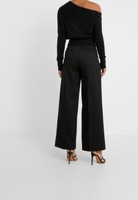 Žena oblečená v černém úzkém svetrnu bez ramen, širokých černých kalhotách a černých sandálech na vysokém podpatku, stojí na bílém pozadí.