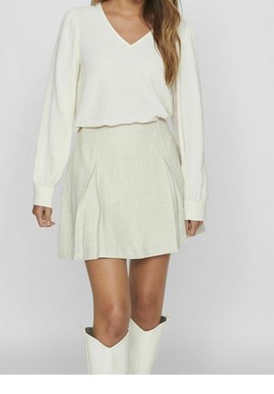 Vrouw met een crèmekleurige blouse met lange mouwen en V-hals, een beige geplooide minirok en witte kniehoge laarzen tegen een eenvoudige lichte achtergrond.