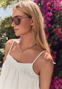 White sleeveless top with gathered neckline; textured fabric. Dainty gold chain necklace with a small charm; brown tinted round sunglasses.