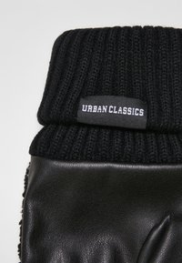 Svarte strikkede hansker med ribbestrikket mansjett, med en glatt skinnhåndflate. En etikett med "URBAN CLASSICS" er sydd på mansjetten.