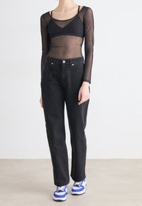 Persona con una blusa negra transparente de manga larga sobre un sujetador negro, jeans negros de cintura alta y zapatillas blancas con detalles en azul sobre un fondo plano.