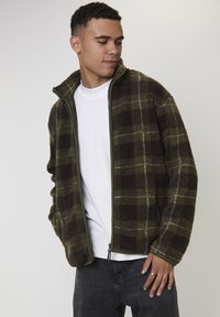 Młody mężczyzna w zielonej i brązowej kratkowanej polarowej kurtce, noszący białą koszulkę i ciemne dżinsy, patrzy w dół i na bok.