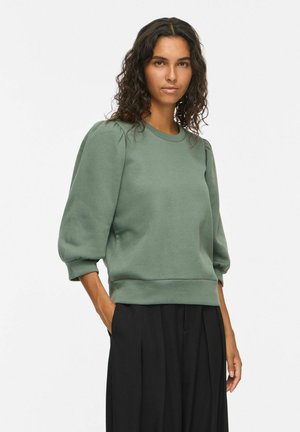 Femme aux cheveux bouclés portant un sweat-shirt vert à manches bouffantes et un pantalon noir à jambes larges, debout avec une main dans la poche.