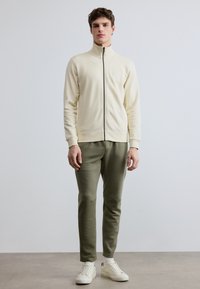 Kermainen vetoketjullinen collegepaita korkealla kauluksella, yhdistetty oliivinvihreisiin kapealöysyyslahkeisiin collegehousuihin, joissa on resorit lahkeissa, ja valkoiset lenkkarit.