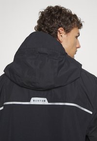 Veste noire avec capuche, présentant une bande réfléchissante et un logo. Tissu texturé avec un design structuré, montrée de dos.