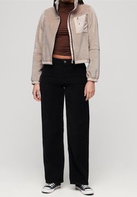 Light beige zip-up jacket with elastic cuffs, brown fitted top, black corduroy wide-leg pants, and black sneakers with white accents.