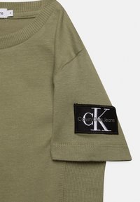 T-shirt in cotone verde oliva con una toppa rettangolare nera con testo bianco sul maniche e colletto a costine.