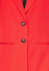 Blazer rouge avec un col cranté, deux boutons noirs et deux poches avant avec des lignes droites et une texture lisse.