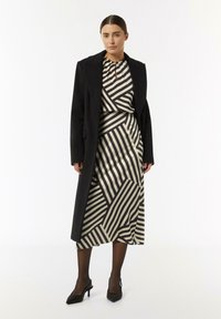Schwarzer Mantel über einem cremefarbenen Kleid mit diagonalen schwarzen Streifen. Das Kleid hat einen gerafften Ausschnitt und ein fließendes Design. Das Modell trägt schwarze Absatzschuhe.