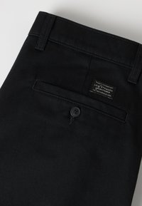 Μαύρο παντελόνι Levi Strauss που δείχνει την πίσω τσέπη με κούμπωμα κουμπιού και μια μικρή ετικέτα μάρκας πάνω από την τσέπη.