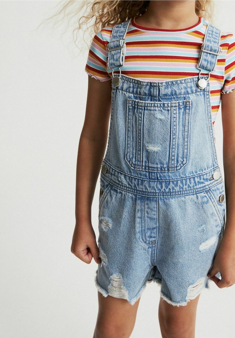 Salopette en denim avec un lavage bleu clair, présentant des détails usés, des bretelles ajustables et une poche avant. Portée avec un t-shirt rayé multicolore.