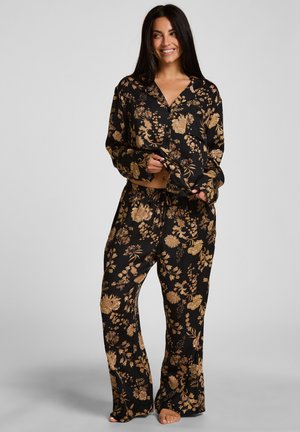 Lächelnde Frau steht barfuß und trägt einen schwarzen Pyjama mit goldenem Blumenmuster, hält den Saum des Hemdes mit einer Hand.