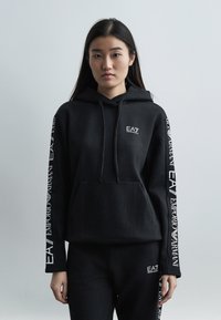 Γυναίκα που φοράει μαύρο φούτερ Emporio Armani EA7 με κουκούλα και αντίστοιχο παντελόνι με λευκά λογότυπα της μάρκας στα μανίκια και στο πόδι του παντελονιού, στέκεται μπροστά σε απλό φόντο.