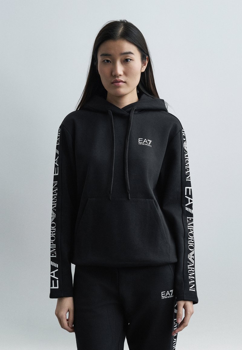 Γυναίκα που φοράει μαύρο φούτερ Emporio Armani EA7 με κουκούλα και αντίστοιχο παντελόνι με λευκά λογότυπα της μάρκας στα μανίκια και στο πόδι του παντελονιού, στέκεται μπροστά σε απλό φόντο.