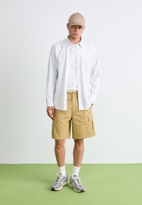 Camisa listrada leve de mangas curtas sobre uma t-shirt branca, combinada com calções de carga bege. Ténis cinzentos com solas brancas e meias.