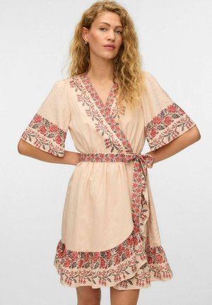 Femme aux cheveux blonds bouclés portant une robe portefeuille beige avec une broderie florale rouge et une ceinture assortie, debout les mains sur les hanches.