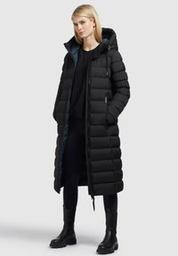 Hosszú fekete pufi kabát kapucnival, steppelt kialakítással, oldalsó zsebekkel és kék bélésével. Fekete bokacsizmával párosítva.